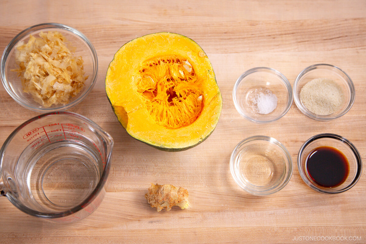 A halved kabocha squash and bowls of water, dried bonito flakes, ginger, salt, sugar, soy sauce, and rice vinegar are arranged on a wooden surface.