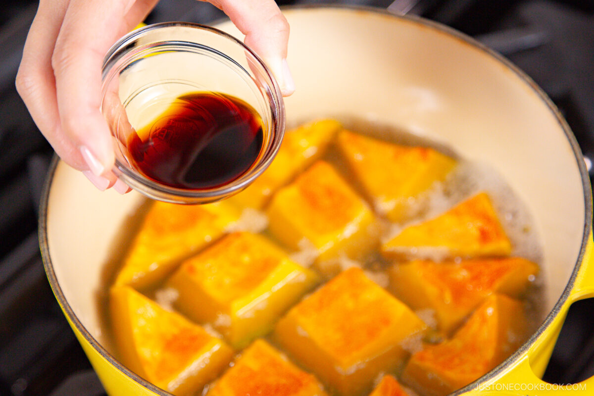 A hand holds a small glass bowl of soy sauce above a pot filled with simmering cubes of bright orange kabocha squash.