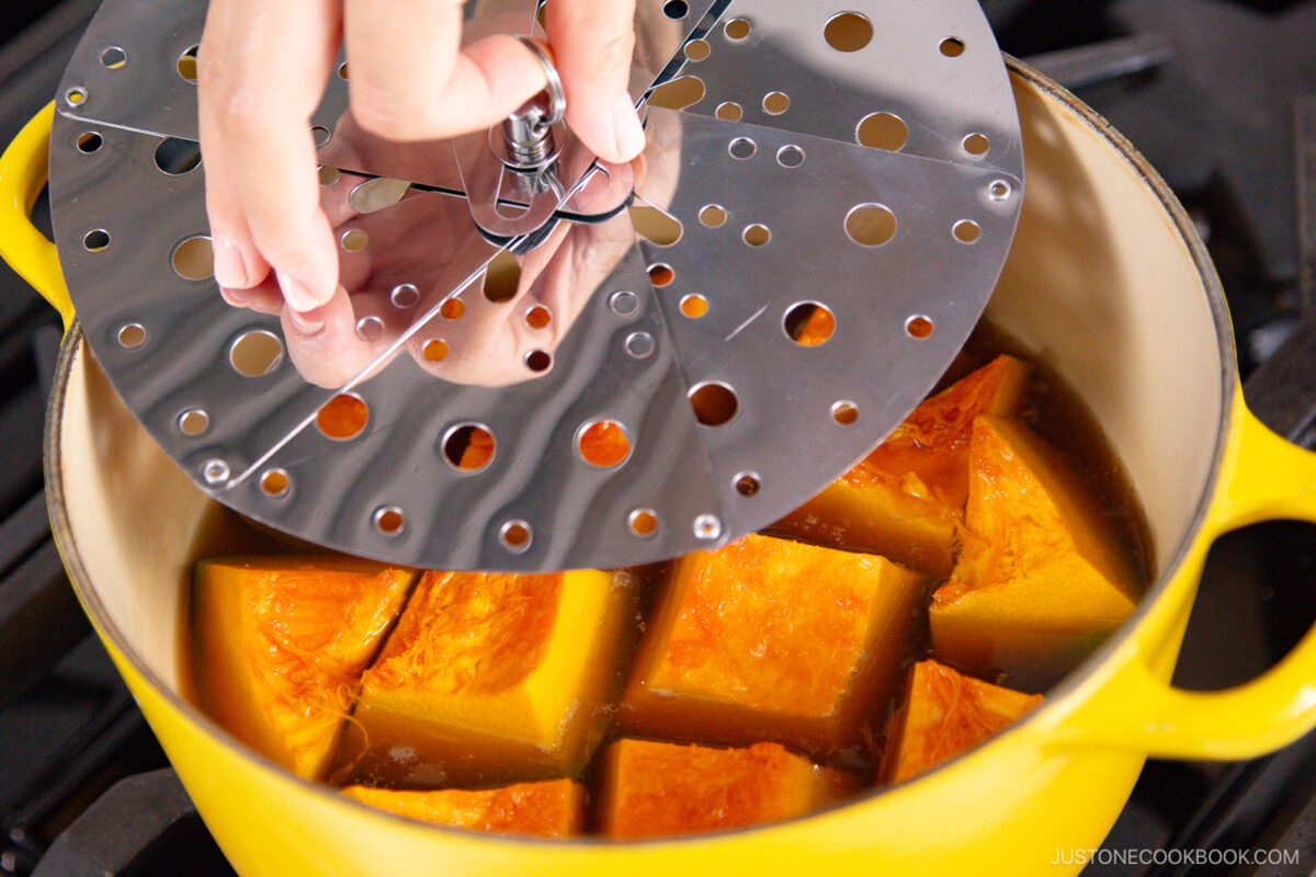 A hand places a metal steamer insert over large chunks of pumpkin in a yellow pot on a stovetop, preparing to steam the vegetables.
