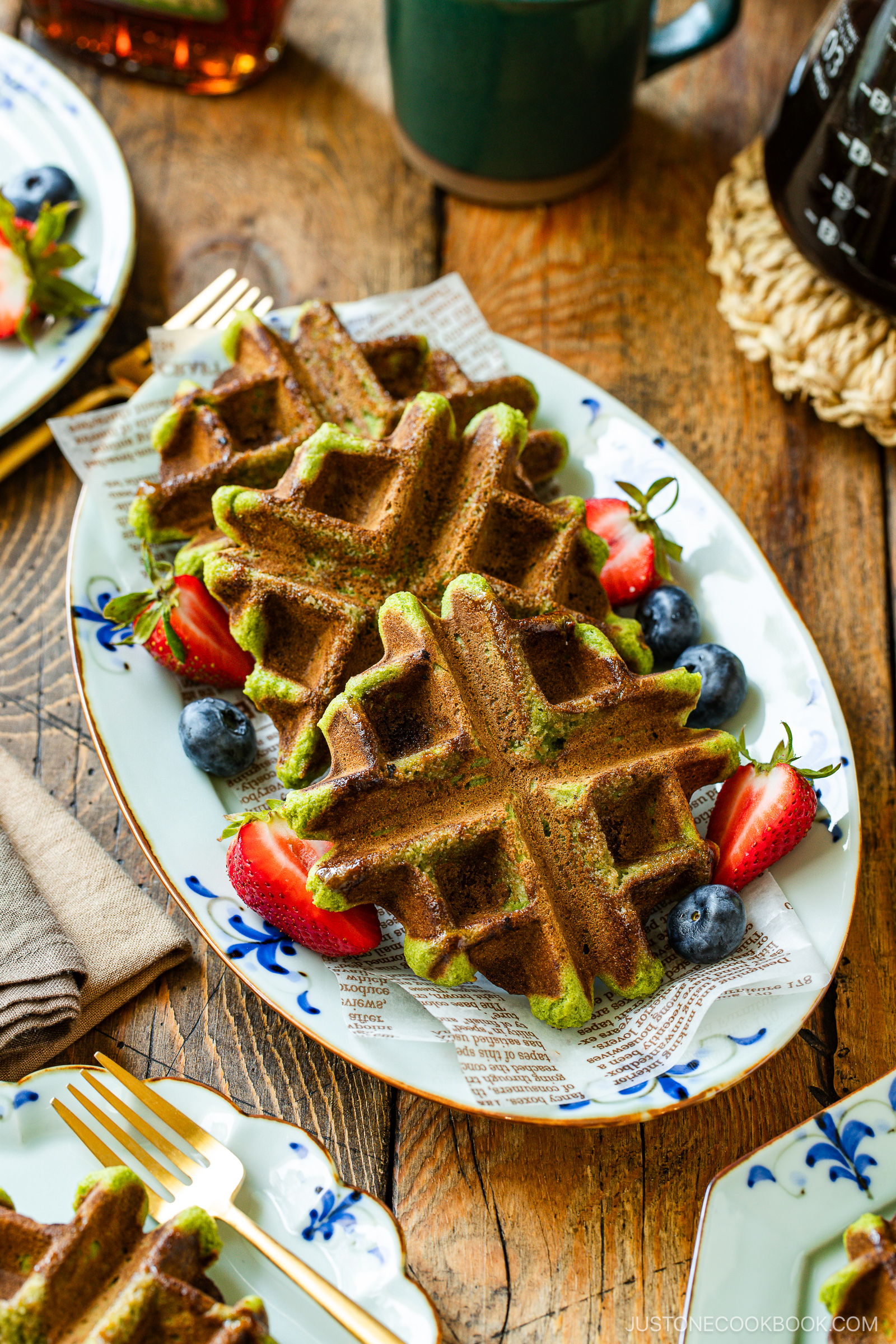 Two green matcha waffles served on a decorative white plate with fresh strawberries and blueberries, set on a rustic wooden table with a cup of coffee in the background.