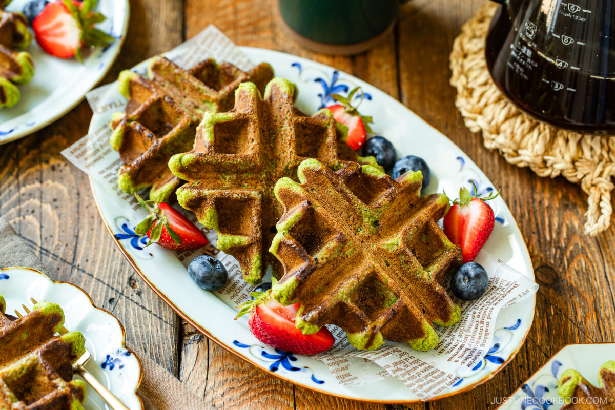 A plate of matcha waffles is served with halved strawberries and blueberries on a rustic wooden table. The waffles are arranged on decorative paper, with coffee and more waffles visible in the background.
