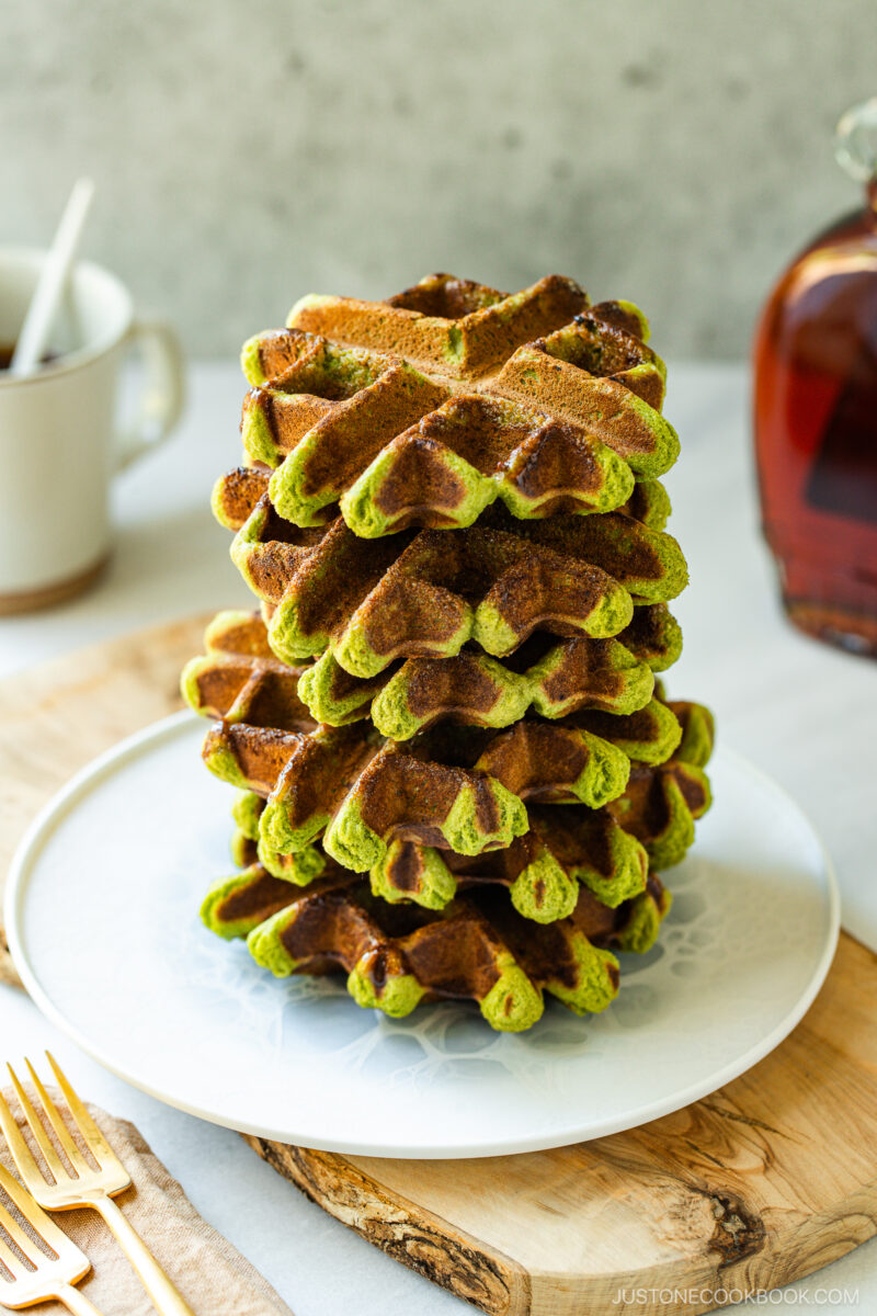 A stack of golden-brown and green matcha waffles is neatly arranged on a white plate, with a cup, syrup bottle, and wooden utensils in the background.
