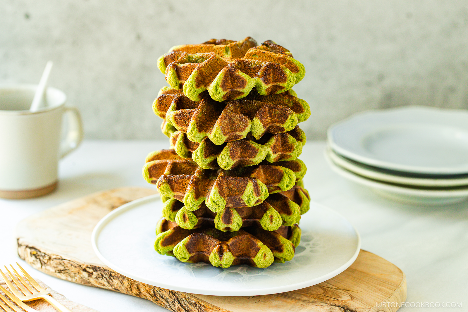 A stack of green and brown matcha waffles sits on a white plate atop a wooden board, with a cup, plates, and forks in the background.
