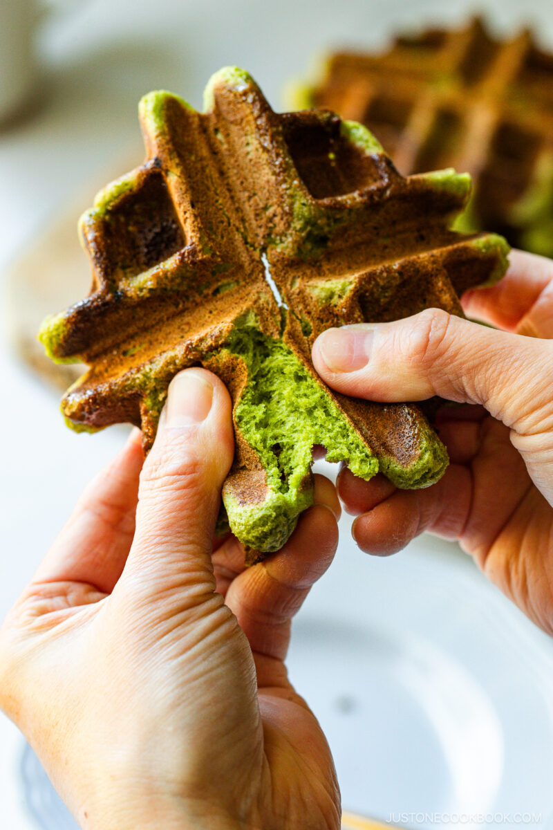 A person pulls apart a fluffy green matcha waffle with their hands, revealing its soft interior. The waffle has a crisp, golden-brown exterior and is shaped with deep square pockets.