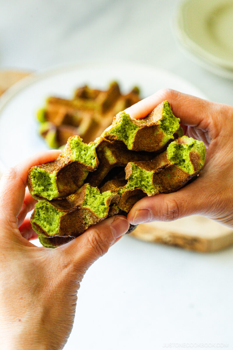 Two hands pull apart a fluffy green matcha waffle, revealing its airy texture. A blurred plate and cutting board are visible in the background.