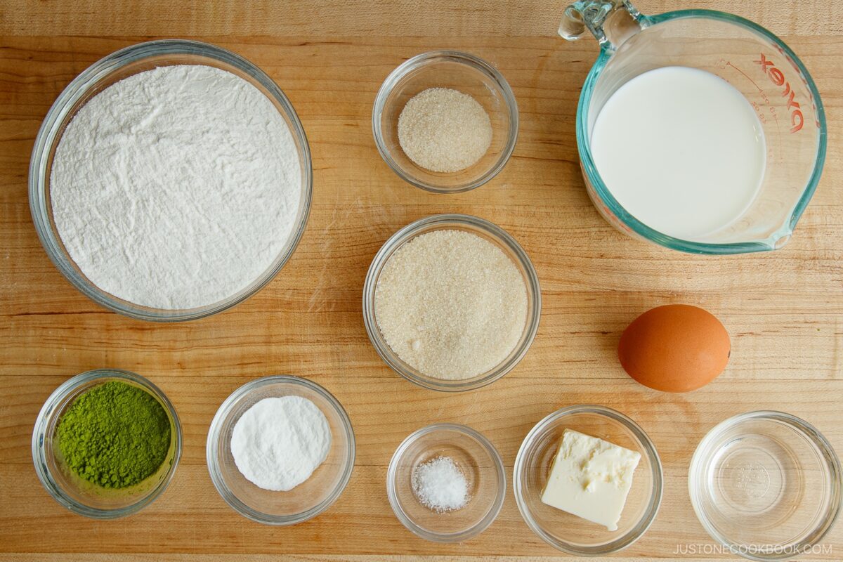 A top-down view of baking ingredients on a wooden surface, including flour, sugar, an egg, milk in a measuring cup, butter, matcha powder, baking powder, salt, and yeast in small glass bowls.