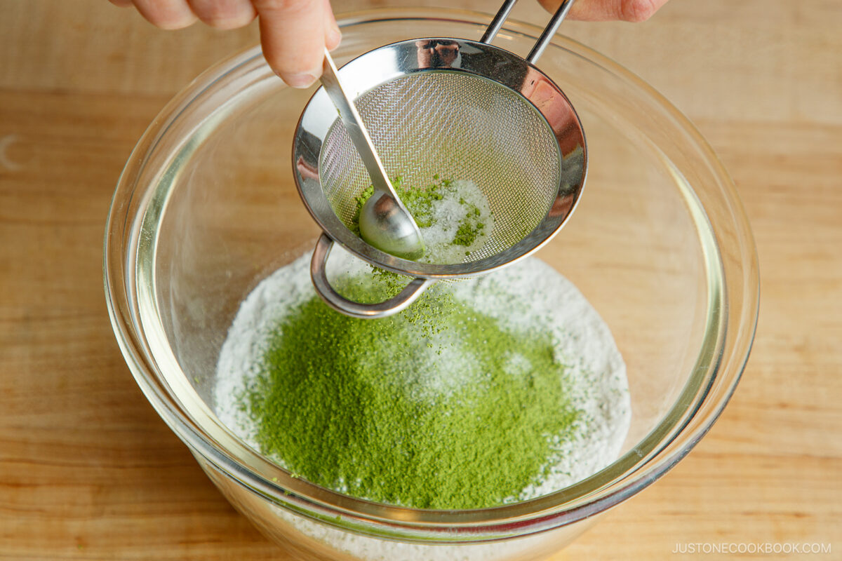 A hand uses a small sifter and spoon to sift green matcha powder into a glass bowl filled with white powdered sugar, all on a wooden surface.