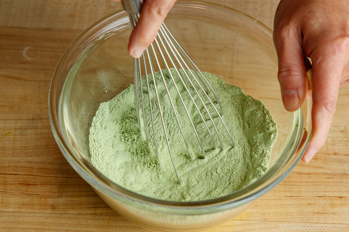 A person whisking light green matcha powder in a glass bowl on a wooden surface.