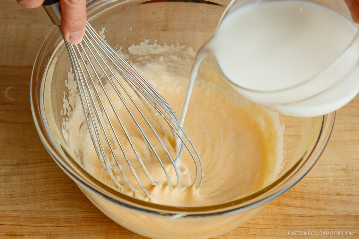 A hand uses a whisk to mix ingredients in a glass bowl while milk is being poured in, all on a wooden surface.