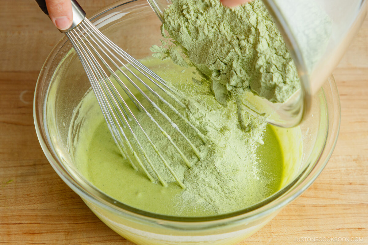 A hand pours green matcha powder into a glass bowl of batter while stirring with a metal whisk on a wooden surface.