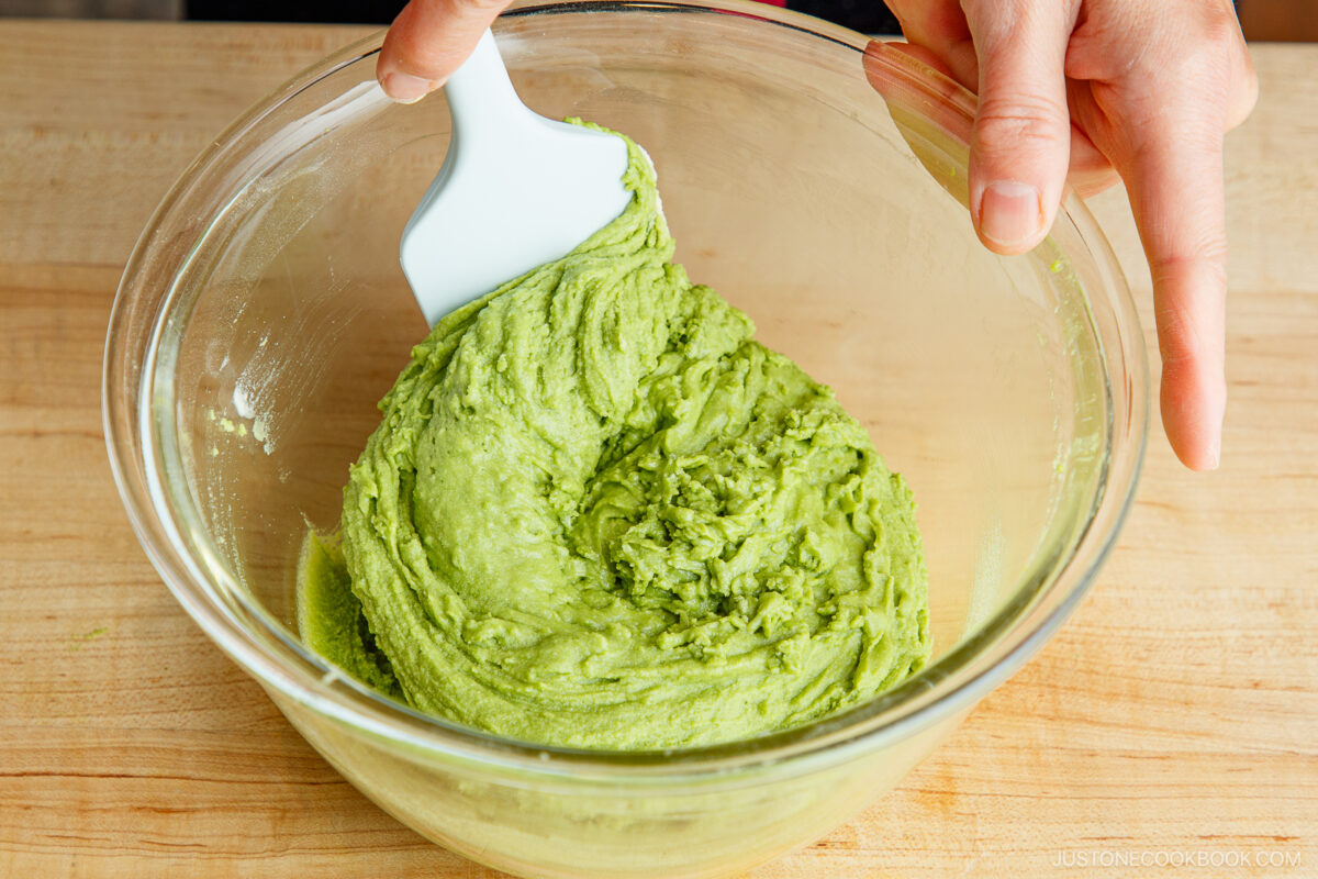 A person uses a white spatula to mix bright green matcha dough in a large clear glass bowl on a wooden surface.