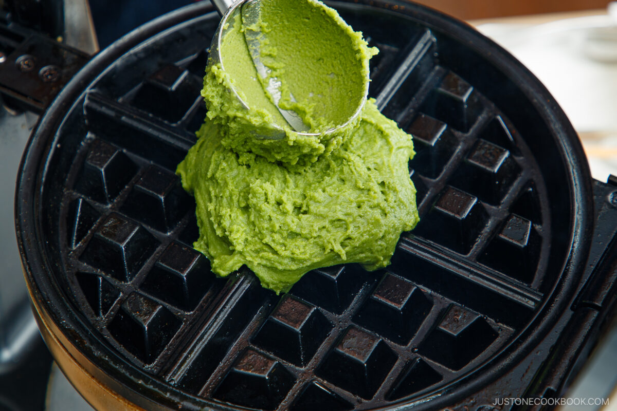 A scoop of green matcha waffle batter being placed onto a preheated waffle iron, ready to be cooked.