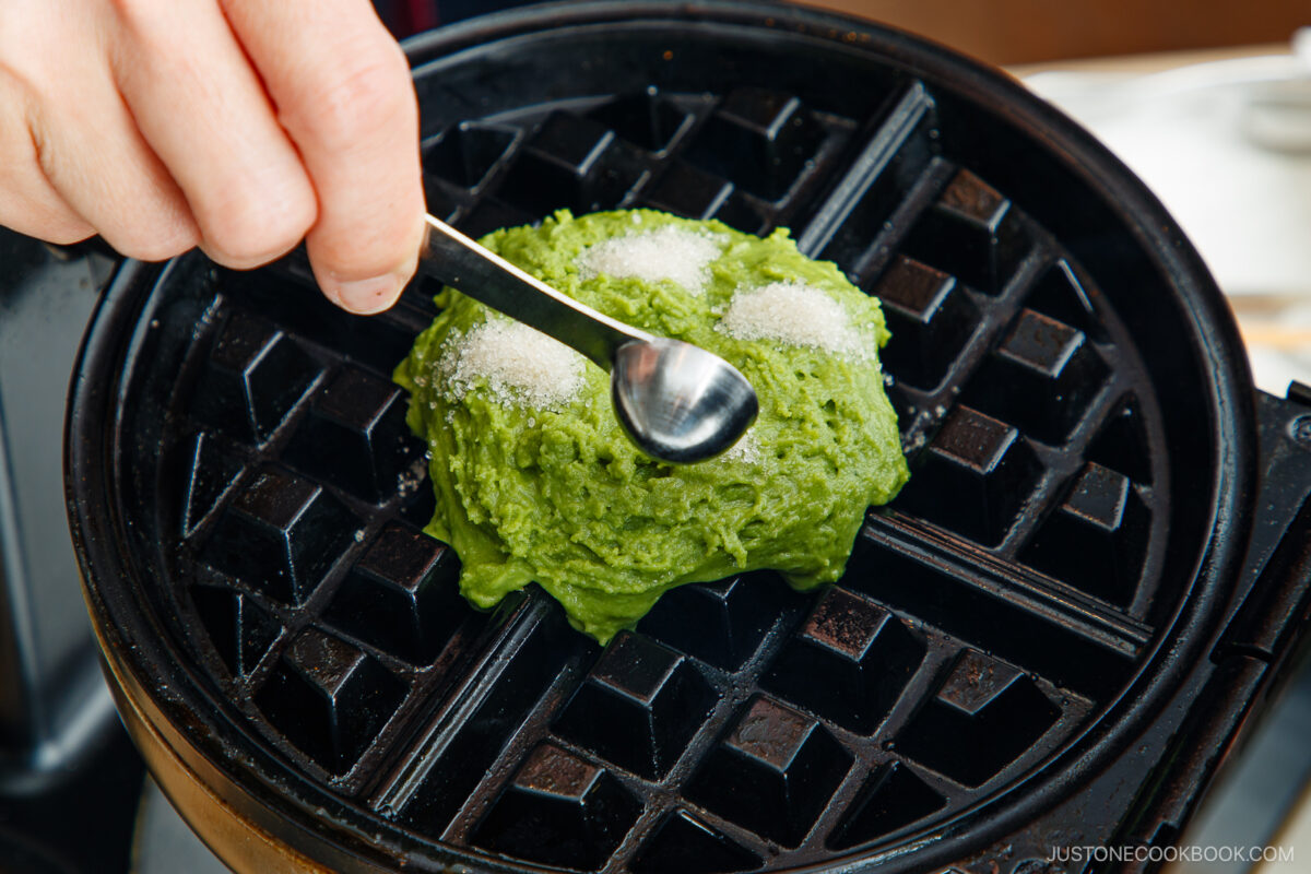 A hand sprinkles sugar with a spoon onto green matcha waffle batter placed on a hot waffle iron, ready to be cooked.