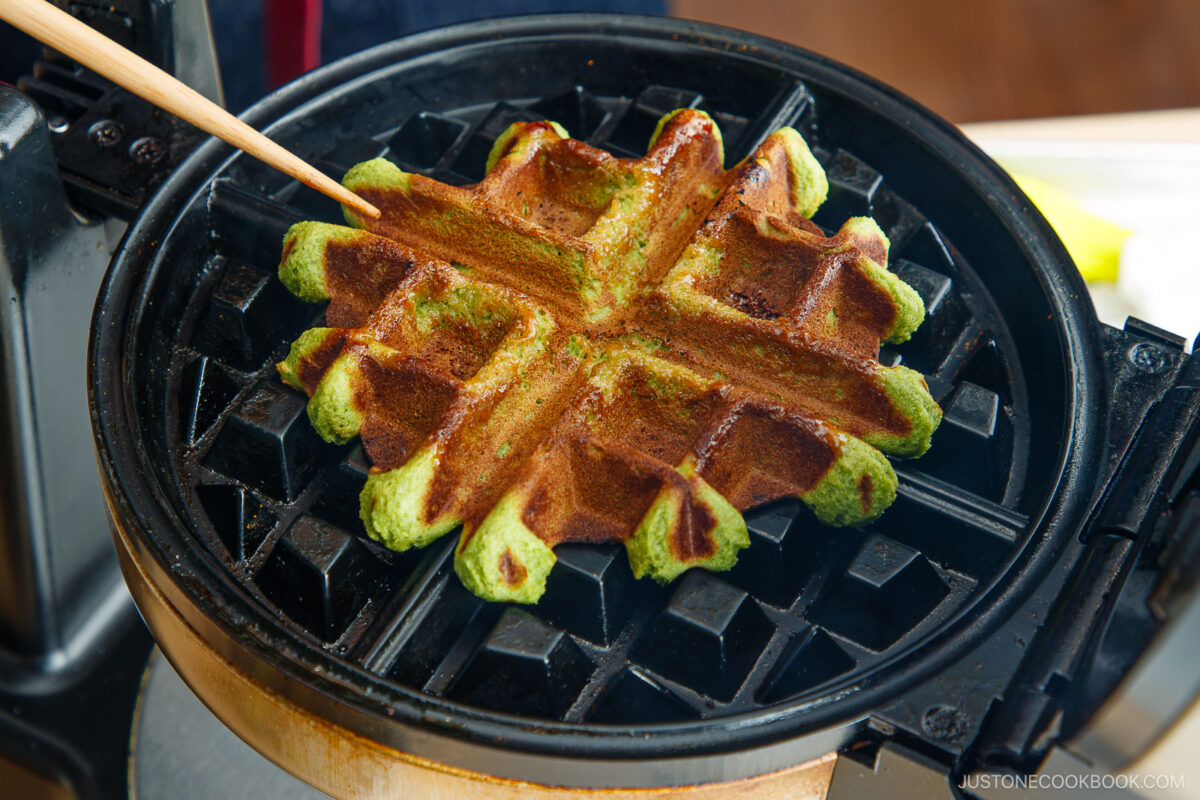 A green matcha waffle is being checked with a skewer while cooking in a round waffle maker.