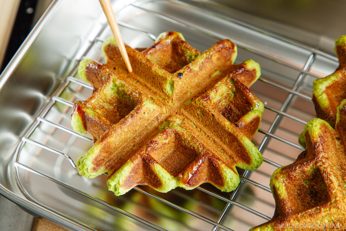 A golden-brown matcha waffle with green edges rests on a wire rack, while a skewer checks its doneness. Another waffle and a metal tray are partially visible in the background.