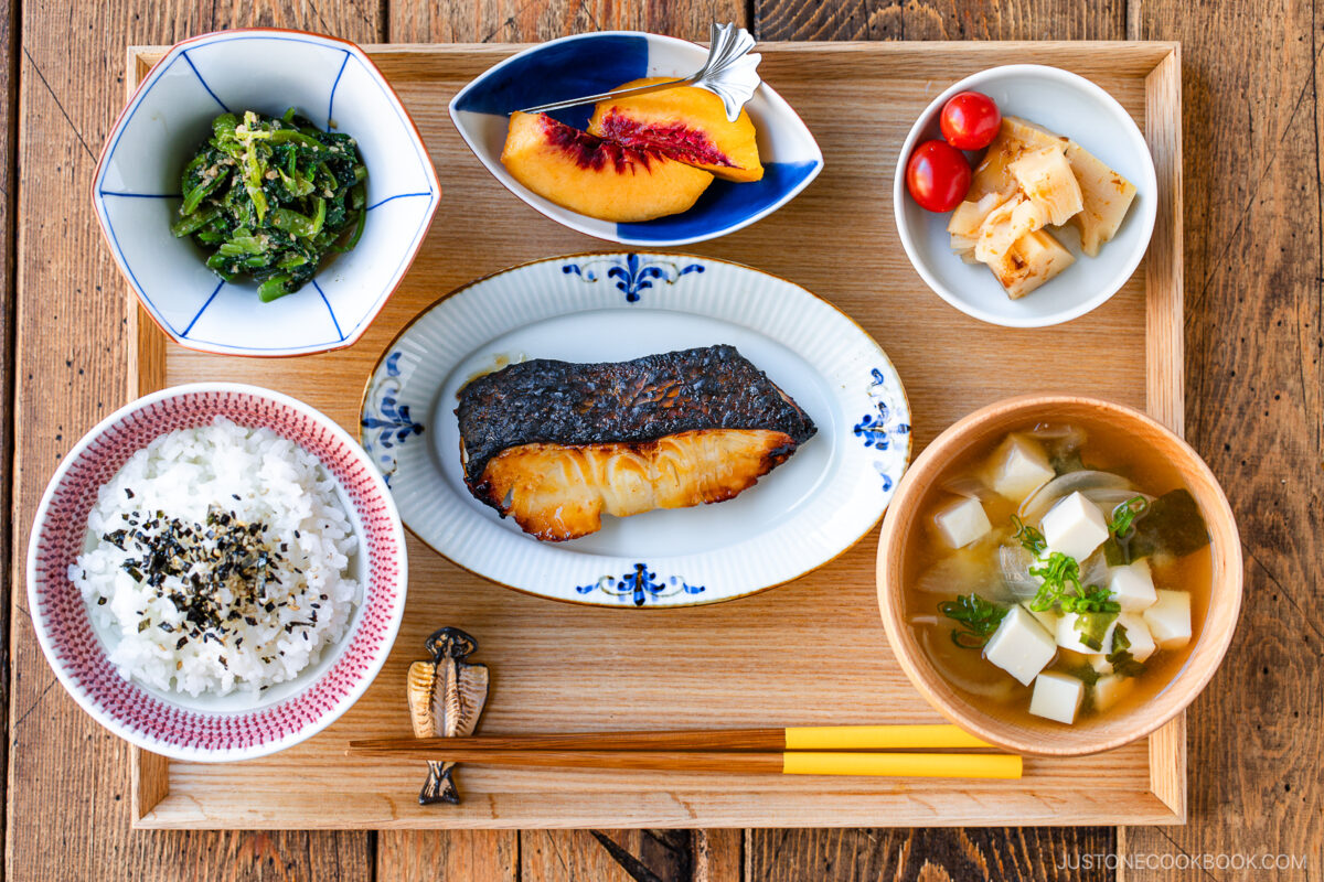 A wooden tray holds a Japanese meal with rice, miso soup with tofu, grilled fish, sautéed greens, pickled vegetables with cherry tomatoes, and a slice of peach, each in separate dishes, with chopsticks on the side.