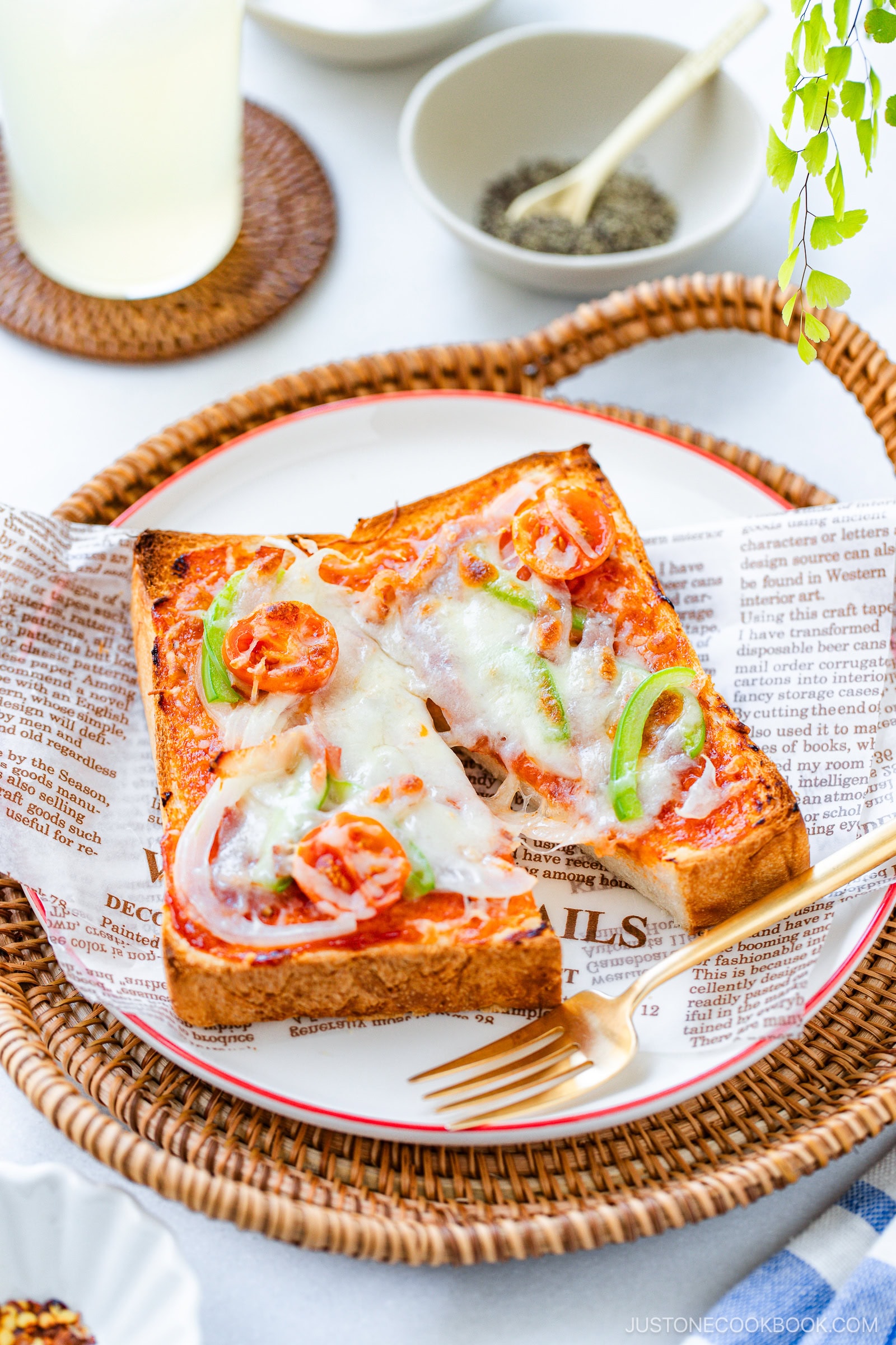 A plate with two slices of thick toast topped with melted cheese, tomatoes, onions, and green peppers. The toast is served on a plate lined with decorative paper, next to a fork, with spices and a drink in the background.