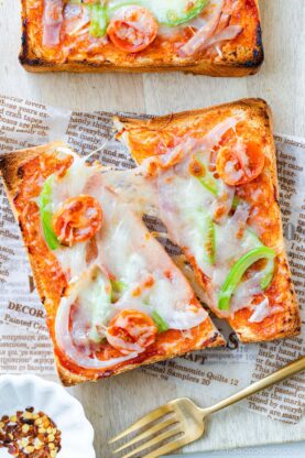 Two slices of toast topped with melted cheese, ham, sliced green bell peppers, onions, and cherry tomatoes, served on parchment paper with a small dish of red pepper flakes and a gold fork nearby.