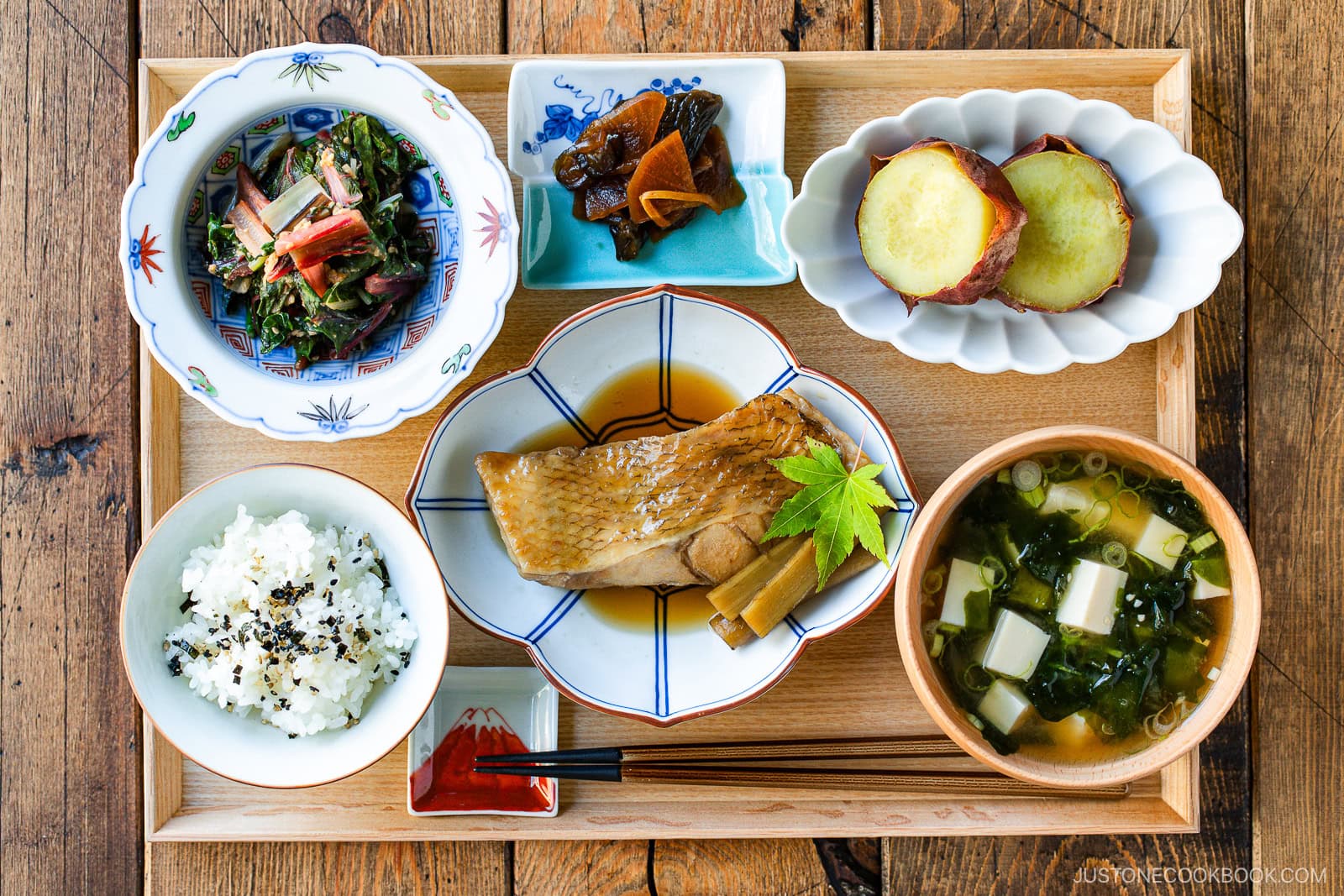 A Japanese meal set on a wooden tray, featuring rice, miso soup, simmered fish, sweet potatoes, pickles, and a spinach salad, each in separate dishes with chopsticks and a small dish of soy sauce.