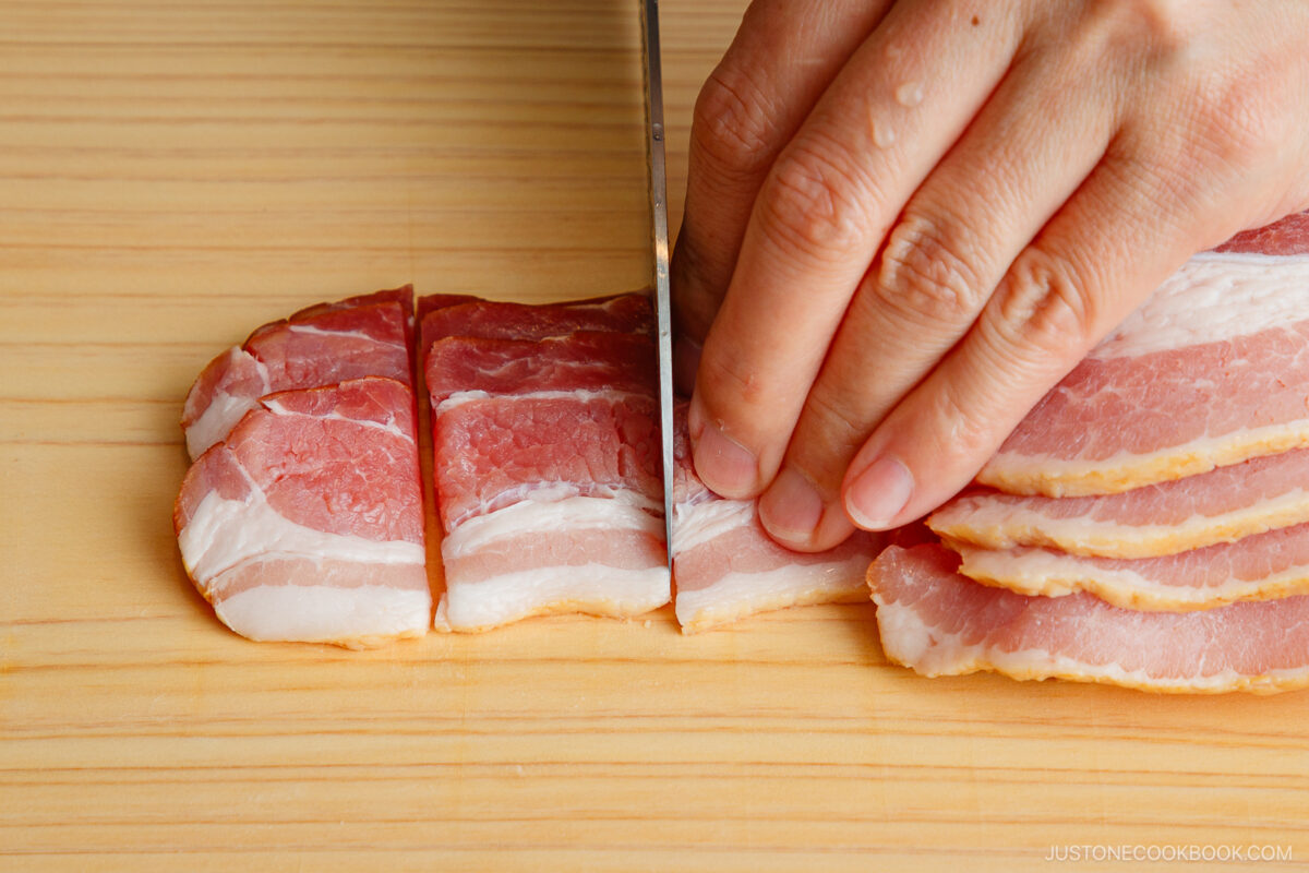 A hand slicing several strips of raw bacon into smaller pieces on a wooden cutting board with a sharp knife.
