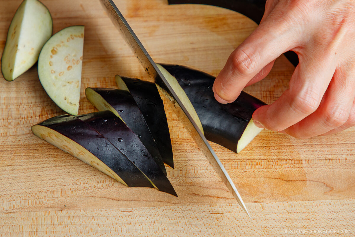 A hand slices an eggplant into thin wedges on a wooden cutting board using a sharp knife.
