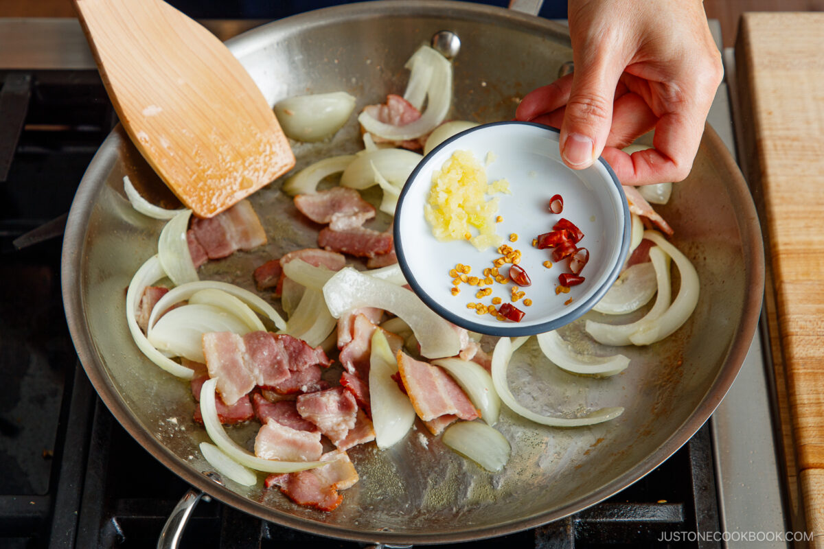 A hand holds a small bowl of minced garlic and sliced red chili over a skillet with sautéing bacon and onion slices, with a wooden spatula stirring the ingredients on a stovetop.