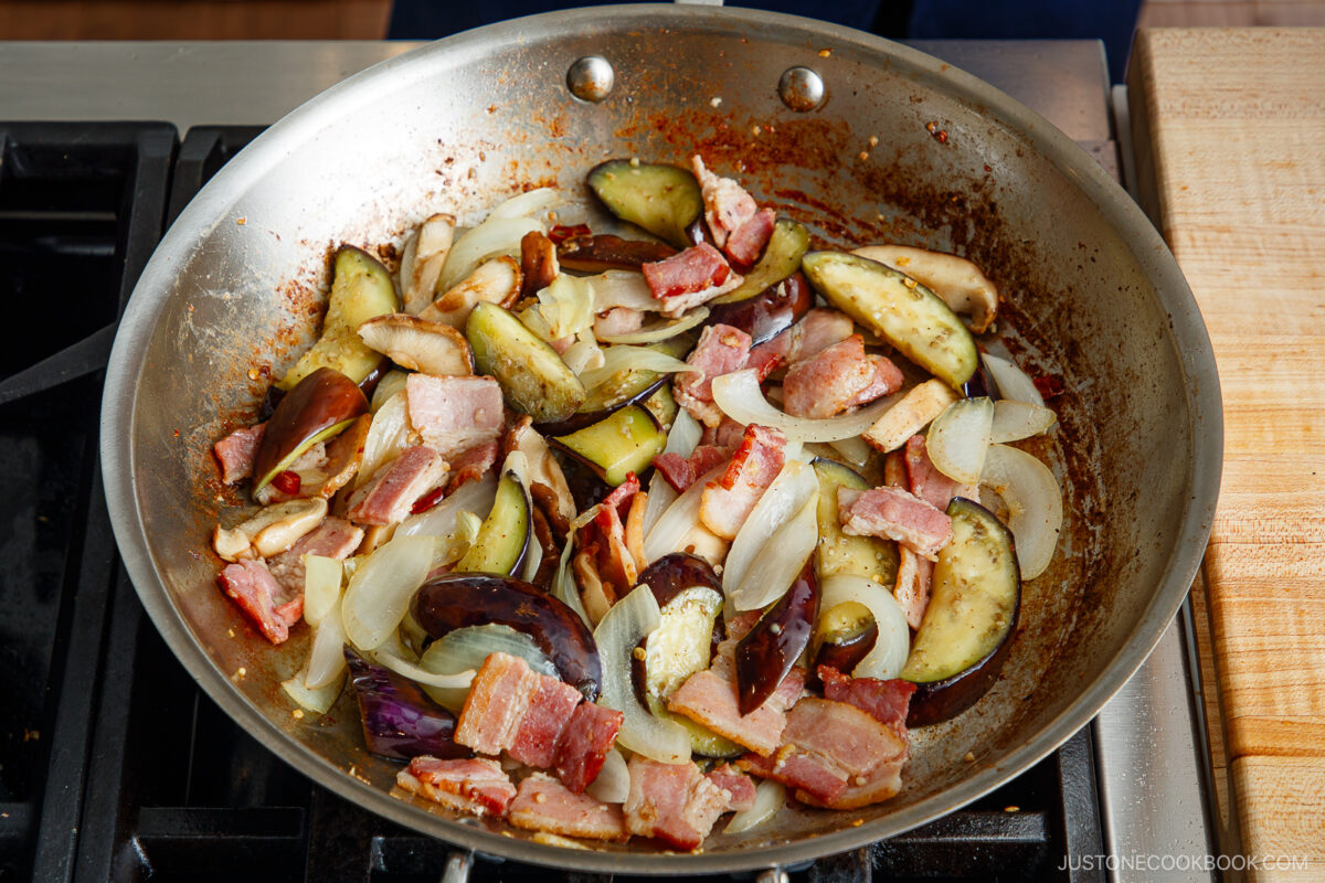 A skillet on a stove contains sliced onions, eggplant, bacon, and mushrooms being sautéed together. The ingredients are lightly browned and mixed, with a wooden cutting board visible nearby.