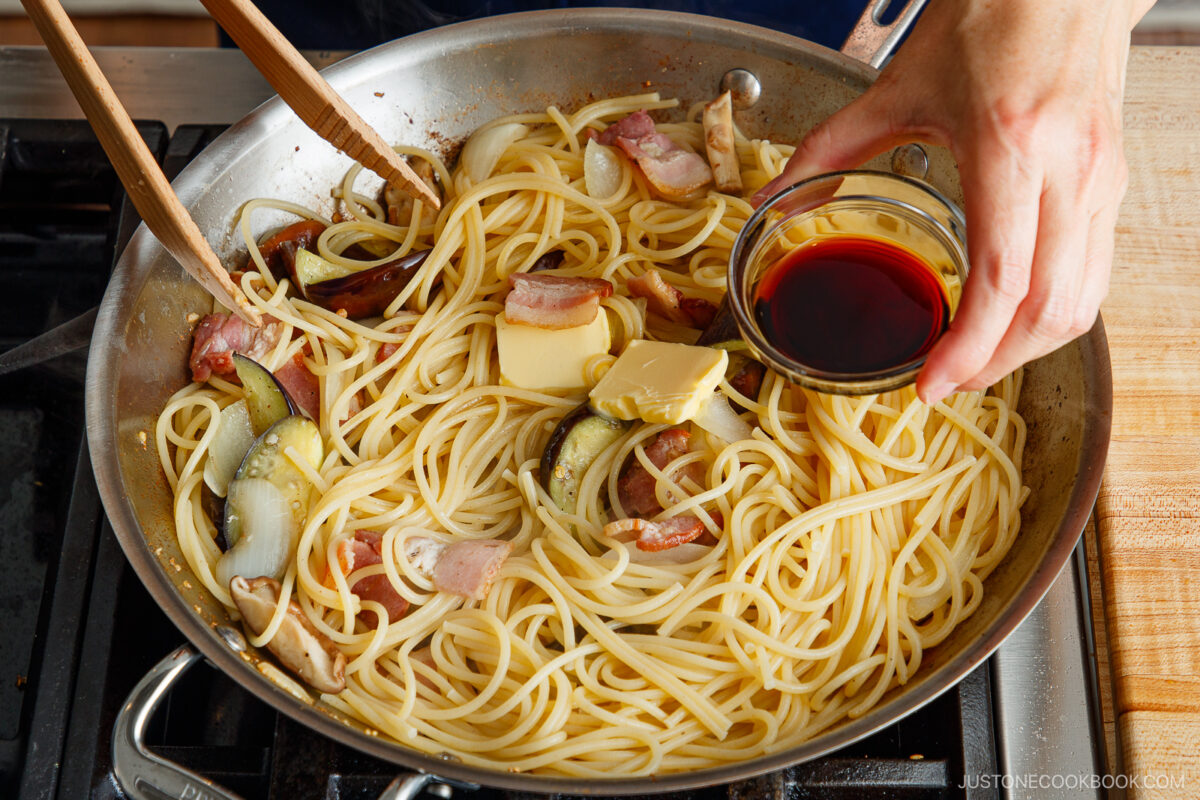 A person adds a small bowl of dark sauce to a skillet of cooked spaghetti with sliced onions, eggplant, bacon, and two pats of butter using wooden tongs on a stovetop.