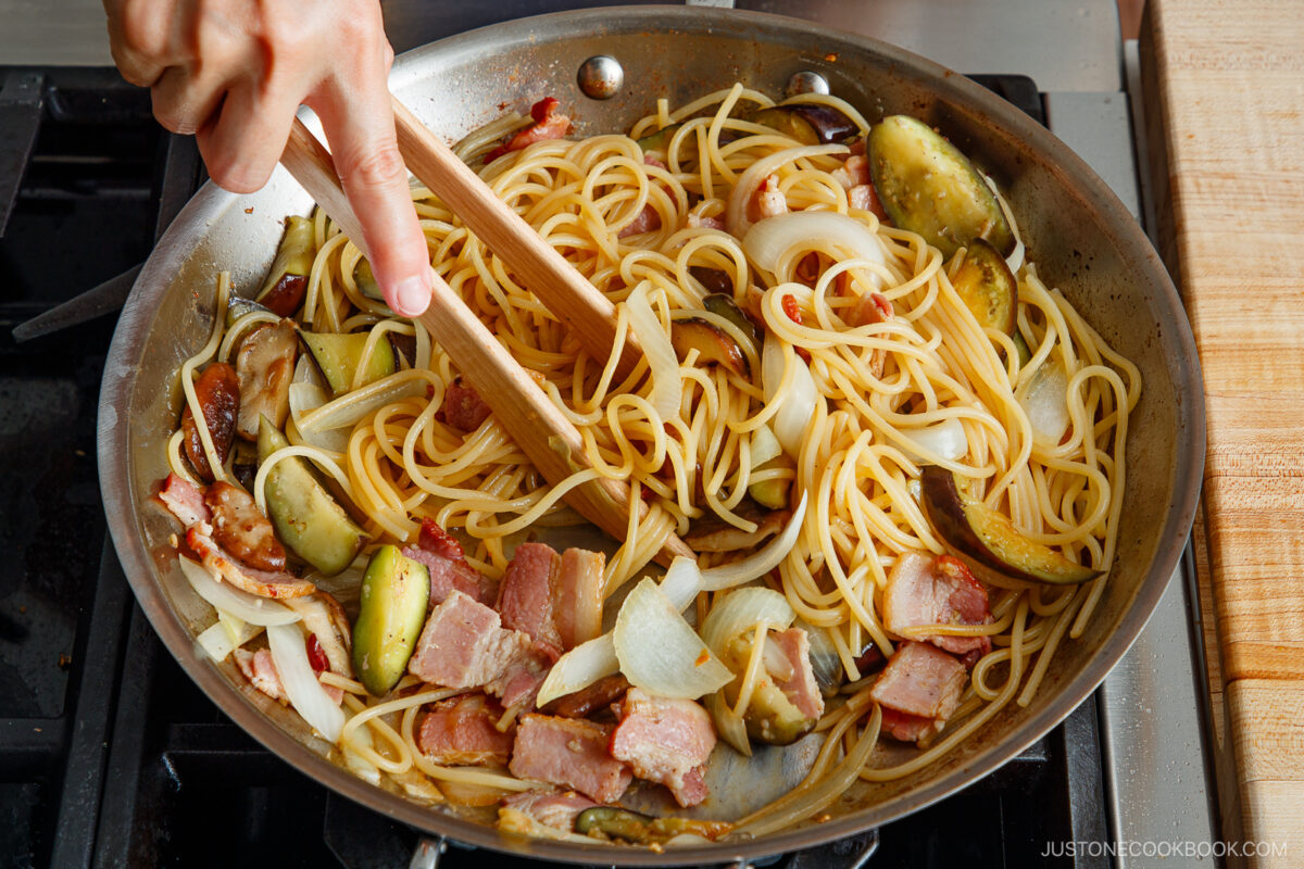 A hand stirs spaghetti, bacon, onion slices, and eggplant in a large skillet on a stove, using wooden utensils.