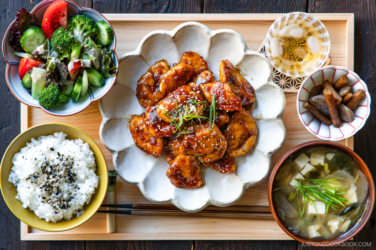A Japanese meal on a wooden tray featuring glazed chicken, white rice with sesame seeds, miso soup with tofu, a fresh salad, simmered root vegetables, and a small dish of pickles.
