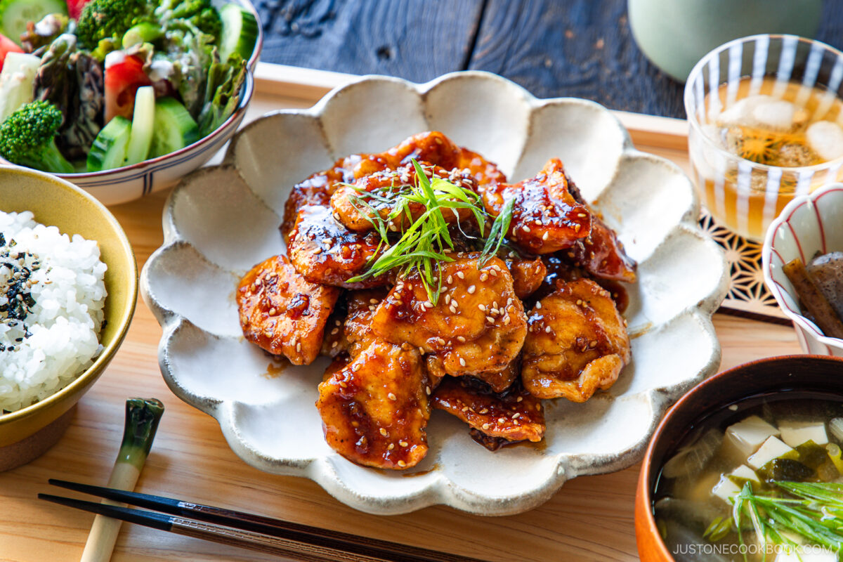 A plate of glazed chicken garnished with green onions and sesame seeds, surrounded by bowls of white rice, soup, and a vegetable salad on a wooden table set for a meal.