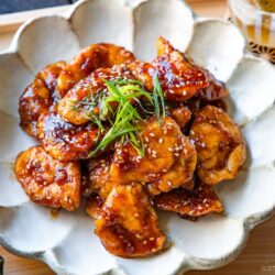 A plate of glazed chicken pieces garnished with sesame seeds and thinly sliced green onions, served on a white scalloped dish with various side dishes around it.