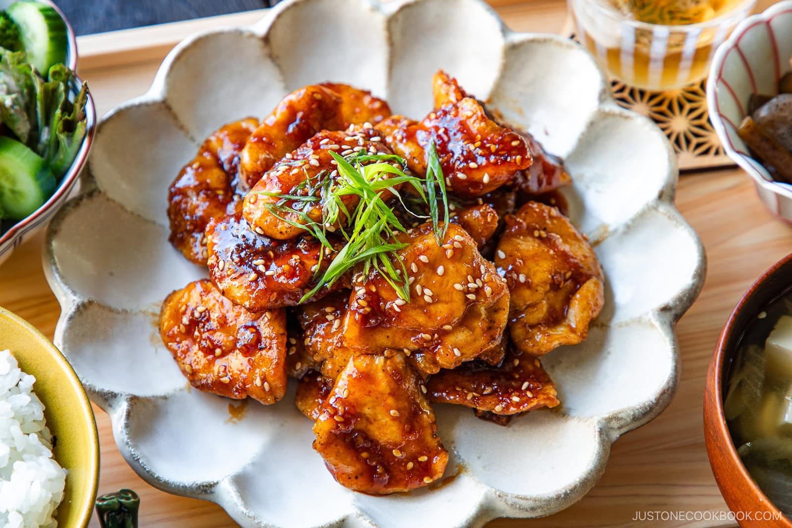 A plate of glazed chicken pieces garnished with sesame seeds and thinly sliced green onions, served on a white scalloped dish with various side dishes around it.