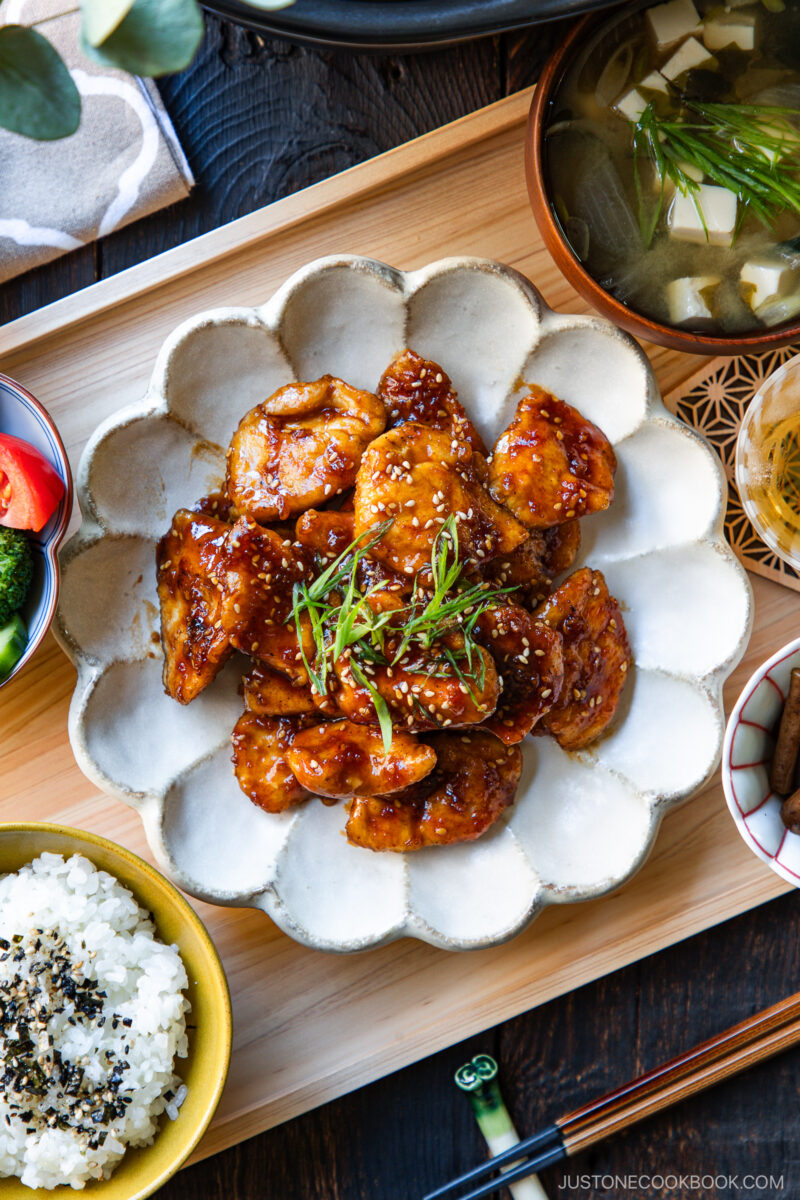 A plate of glazed chicken garnished with green onions is served with rice, soup with tofu, and assorted side dishes on a wooden tray, presented in a Japanese-style meal.