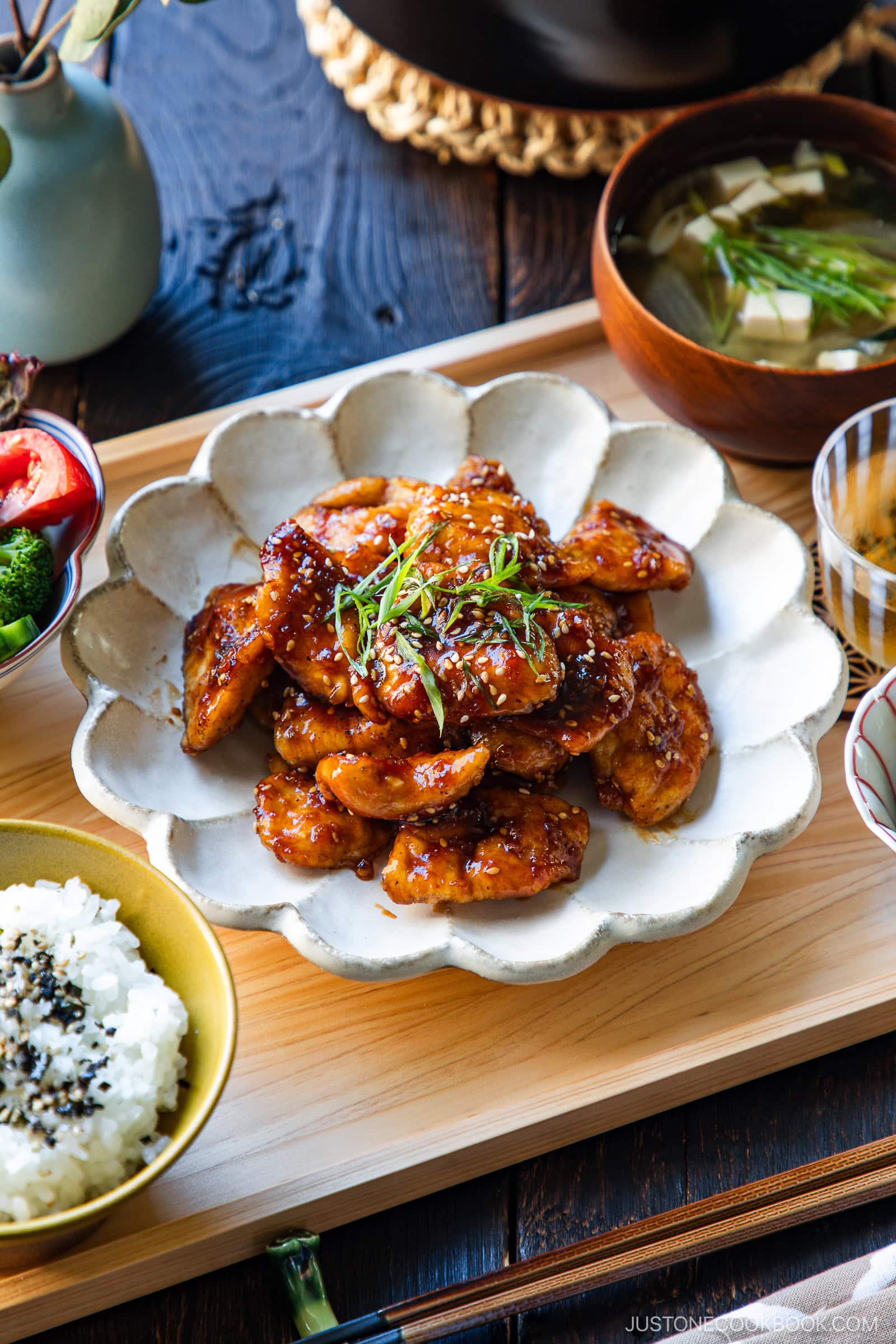 A plate of glazed chicken garnished with green onions and sesame seeds, served with bowls of steamed rice, soup, salad, and side vegetables on a wooden tray.