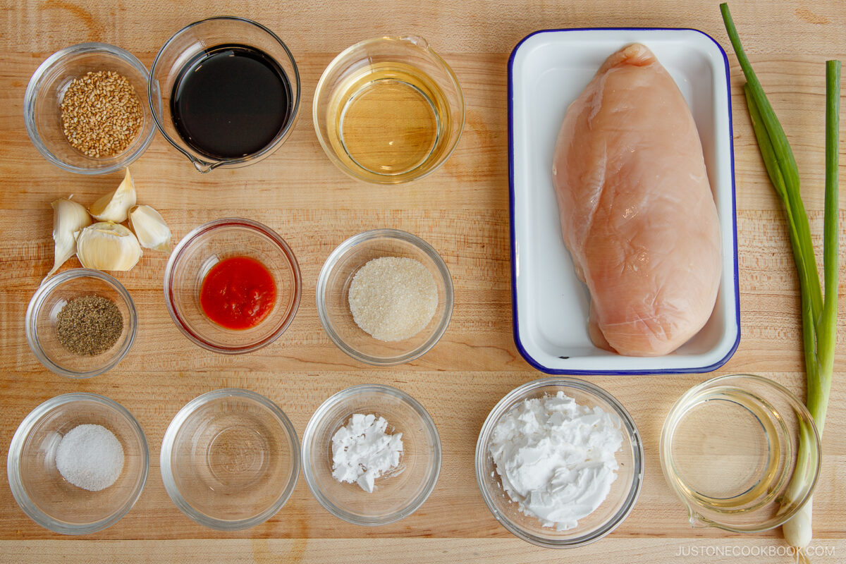 Top-down view of ingredients on a wooden surface, including a raw chicken breast, various sauces, seasonings, garlic cloves, cornstarch, coconut milk, green onions, and small bowls of spices and liquids.
