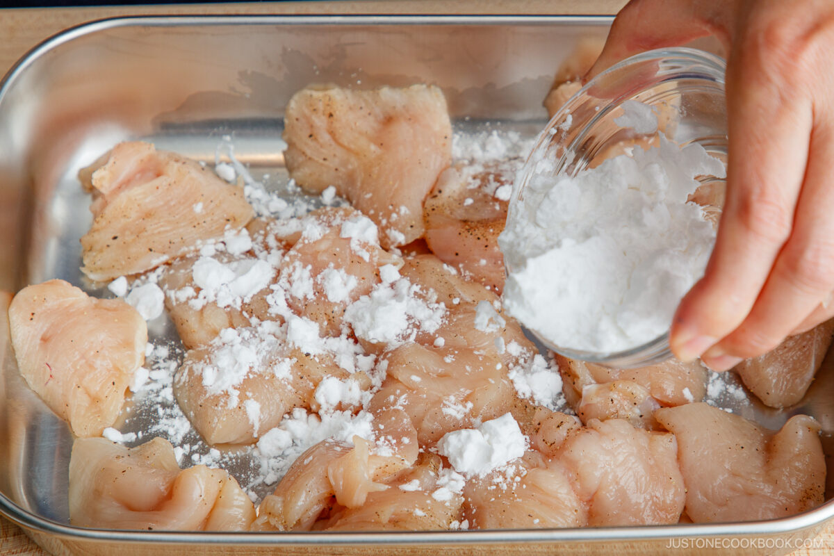 Raw chicken pieces in a metal tray are being sprinkled with a white powder, likely cornstarch, from a small glass bowl by a hand.