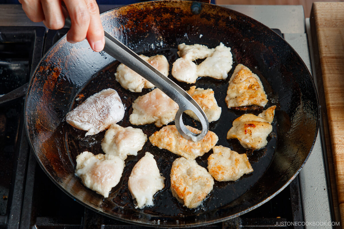 A hand holds metal tongs, turning pieces of chicken frying in a pan on a stovetop. The chicken pieces are both uncooked and golden brown, showing different cooking stages.