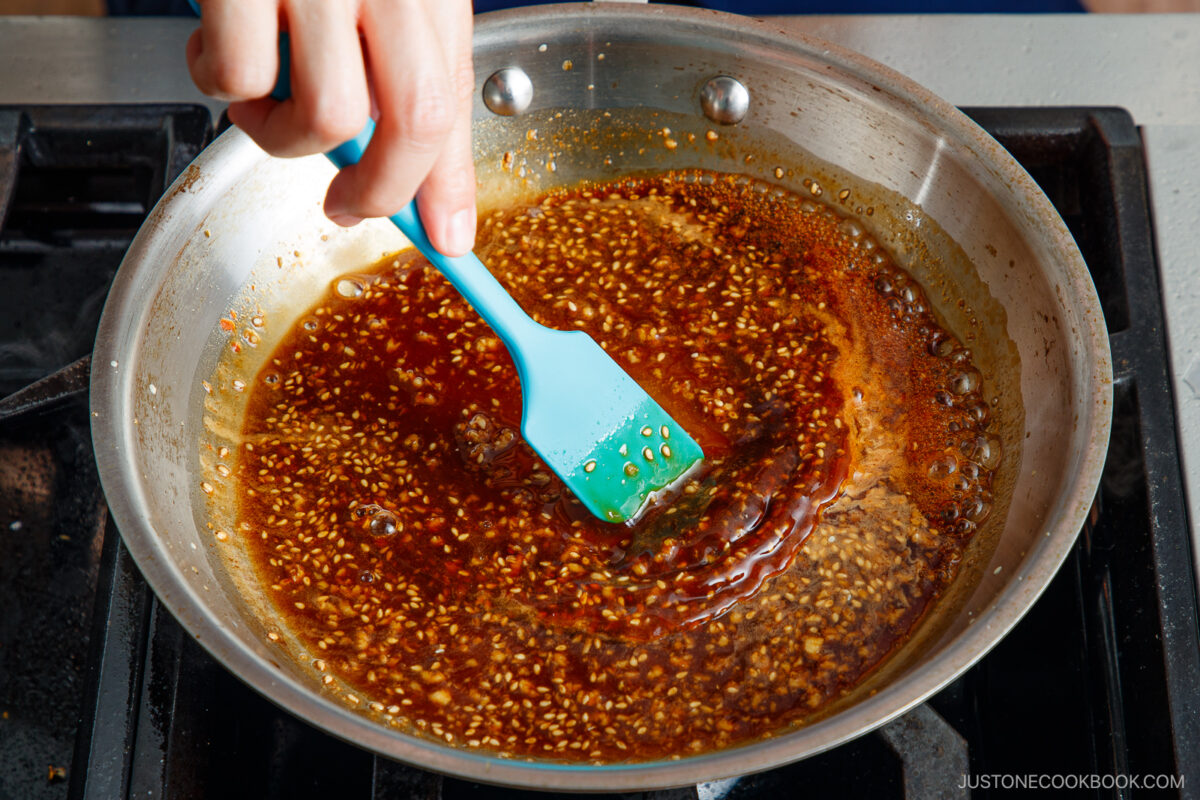 A hand stirs a pan of bubbling brown sauce with sesame seeds using a blue spatula on a stovetop.
