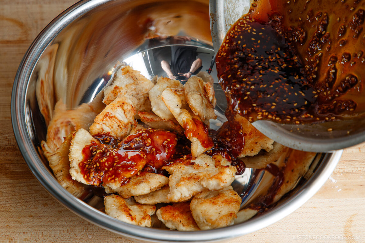 Grilled chicken pieces in a metal bowl are being topped with a dark, glossy sauce containing sesame seeds, poured from another bowl on a wooden surface.