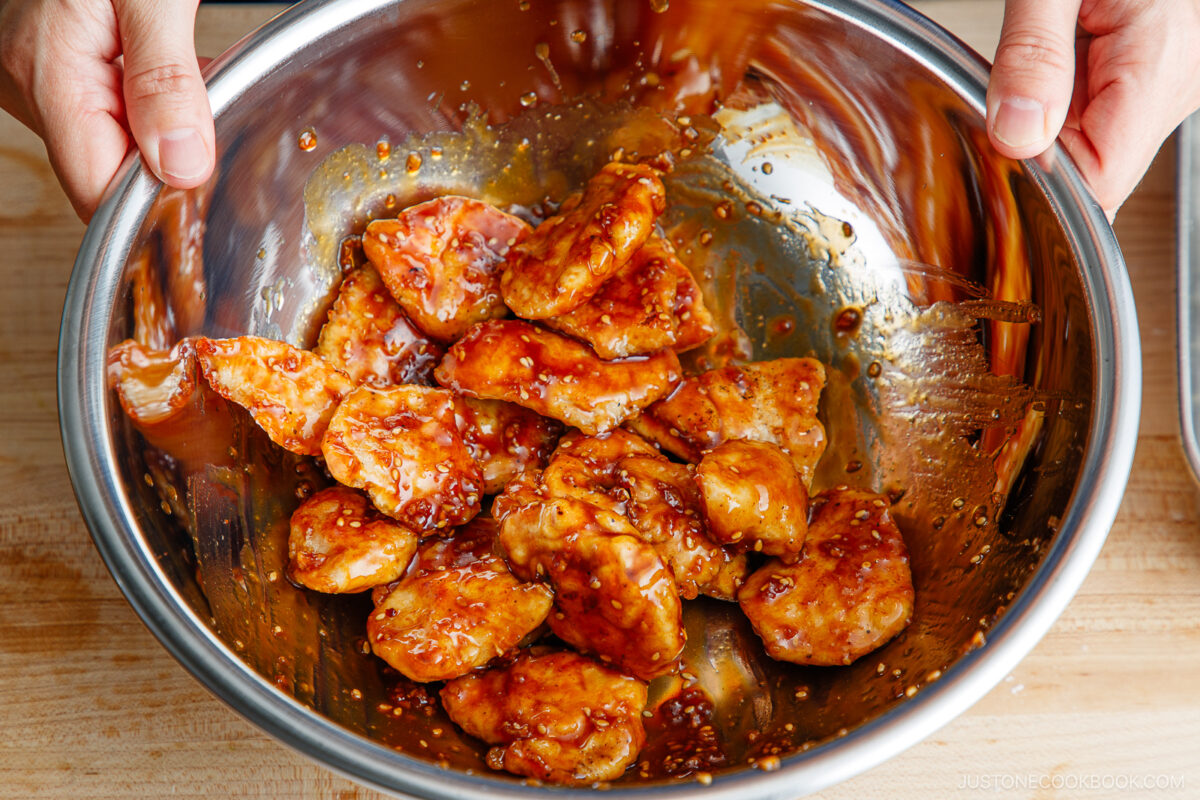 A close-up of hands mixing crispy chicken pieces coated in a shiny brown sauce inside a large stainless steel bowl on a wooden surface.