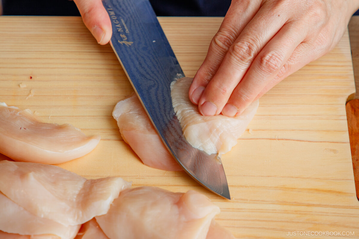 A person slices raw chicken breast into thin pieces on a wooden cutting board using a sharp kitchen knife.