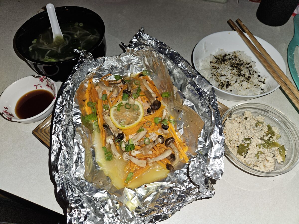 A meal with baked fish and vegetables in foil, a bowl of soup with a spoon, a dish of white rice topped with seaweed, a small bowl of ground meat with green pepper, and a dipping sauce on a countertop.
