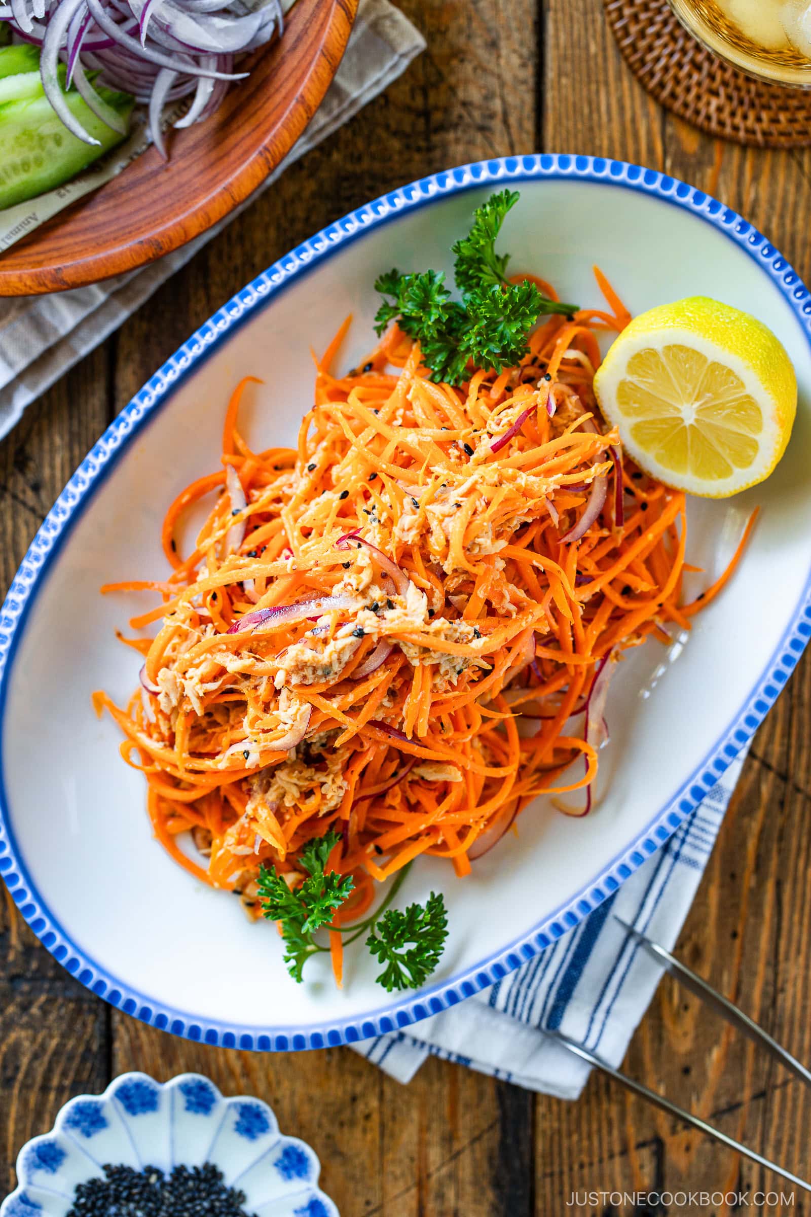 A white oval dish with shredded carrot and chicken salad, garnished with parsley and a lemon wedge, sits on a wooden table. Sliced vegetables and a bowl of black sesame seeds are visible nearby.