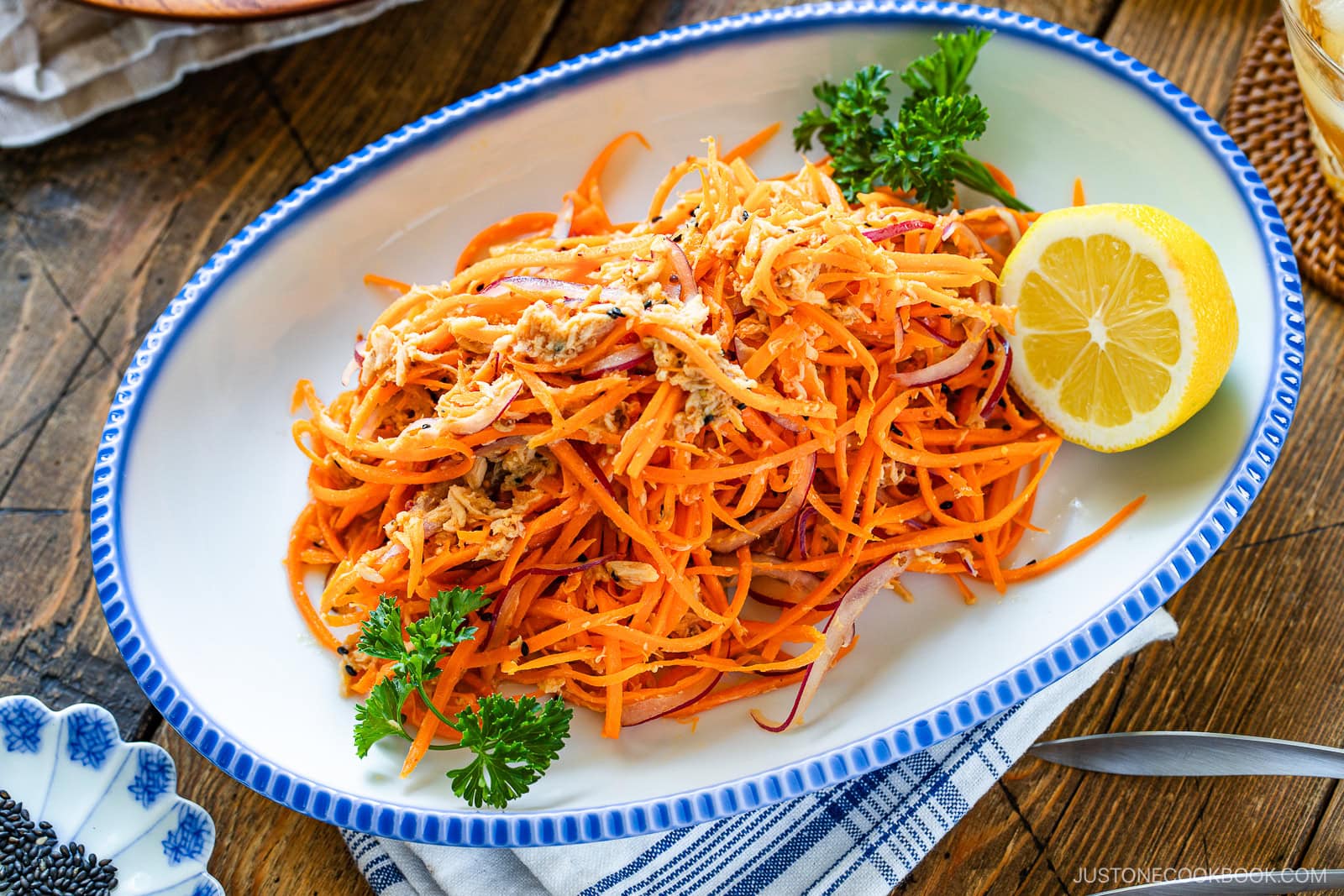 A white oval plate with a blue rim holds a salad made of shredded carrots and what appears to be tuna, garnished with parsley and served with a lemon half on the side. The plate is on a wooden table.