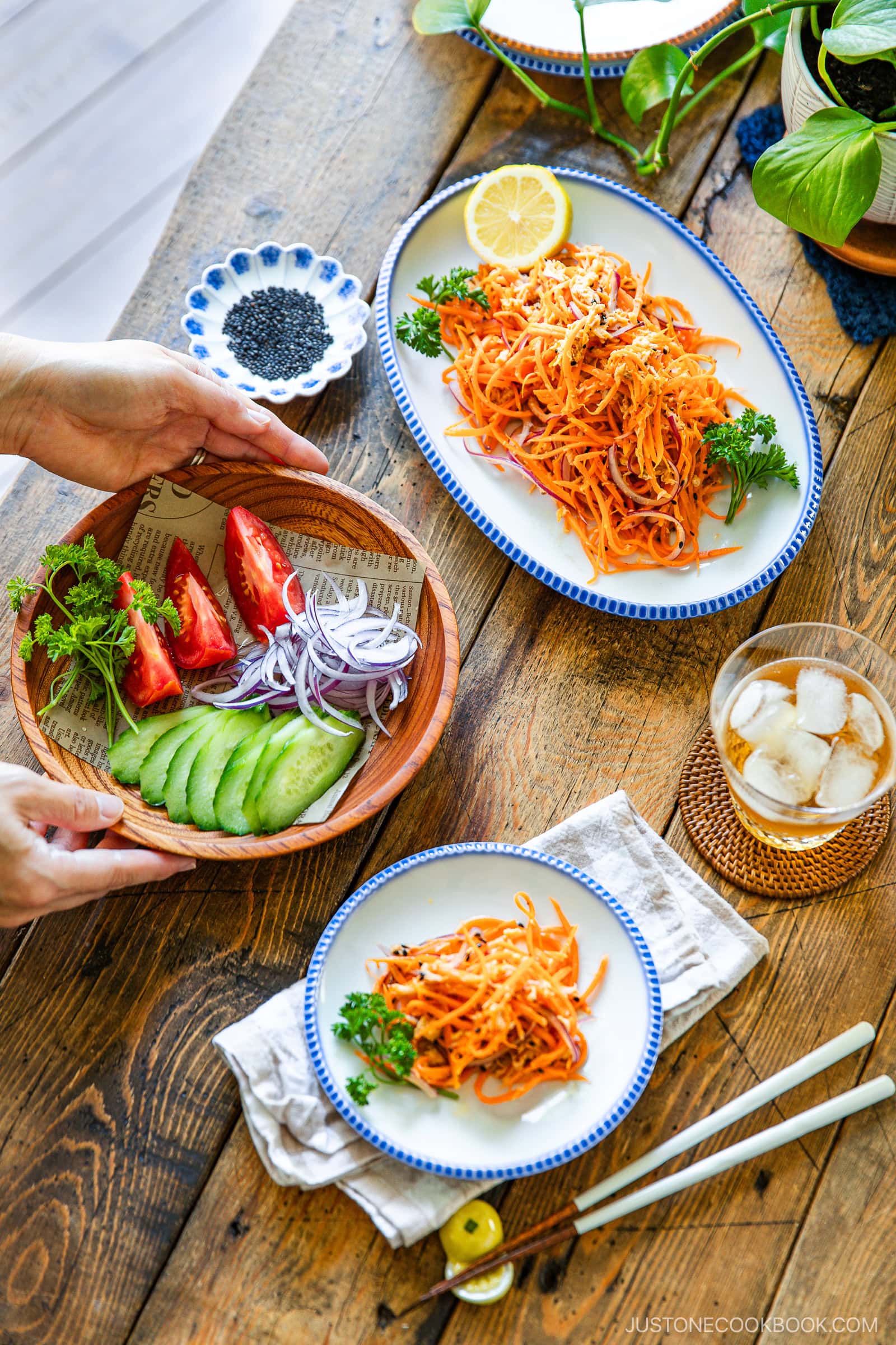 A wooden table set with plates of shredded carrot salad, sliced cucumbers, tomatoes, red onions, a dish of black sesame seeds, and a glass of iced drink. Hands are serving a bowl of vegetables.