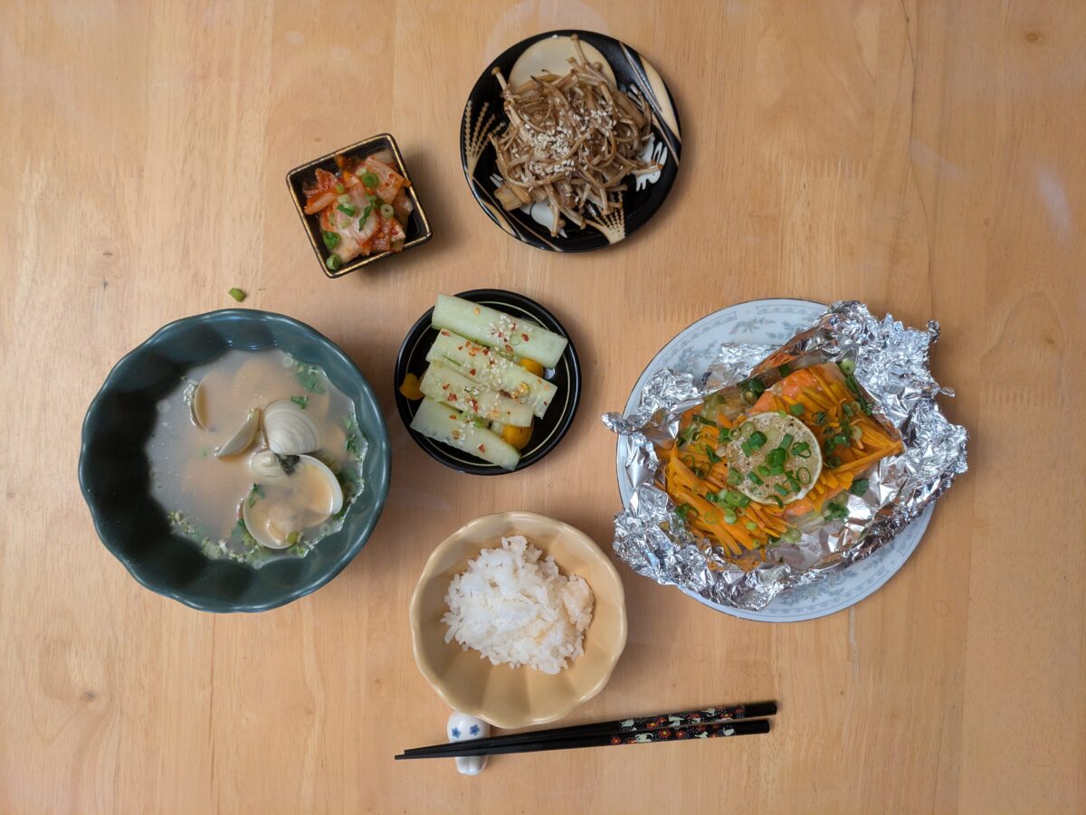 A Japanese meal on a wooden table with clam soup, rice, cucumber salad, kimchi, mushroom salad, and a foil-wrapped steamed fish dish. Black chopsticks rest near the plates.