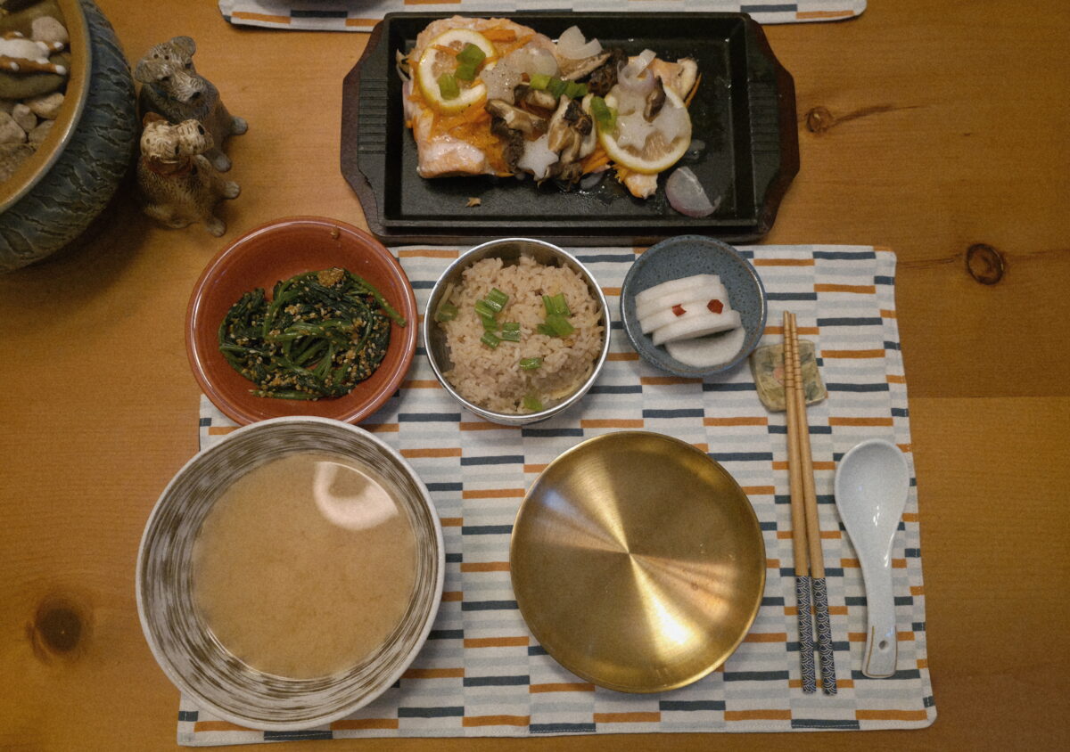 A Korean meal set on a wooden table with miso soup, seasoned spinach, rice with scallions, a metal lid, pickled radish, grilled fish with vegetables, chopsticks, and a spoon on a patterned placemat.