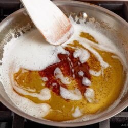A stainless steel pan on a stove contains bubbling brown butter, being stirred with a white spatula; some brown bits are visible in the golden liquid.