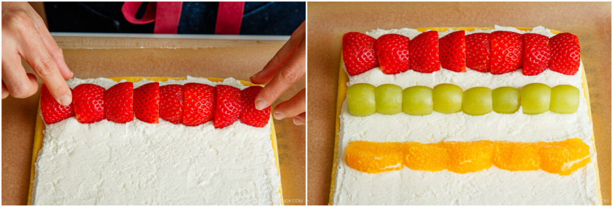 A person arranges halved strawberries on a rectangular cake covered with whipped cream, next to rows of green grapes and orange slices.